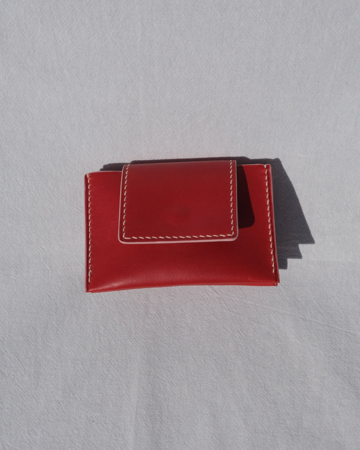 A small red leather card holder or wallet with white contrast stitching, photographed on a light gray textured background with a soft shadow.